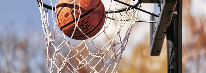 An orange basketball going through a white netted hoop with a black backboard against a blue and cloudy sky.