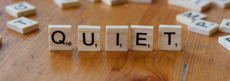 A selection of Scrabble titles that spell out 'Quiet' on a light wooden surface as well as other random Scrabble title on the back of the table.